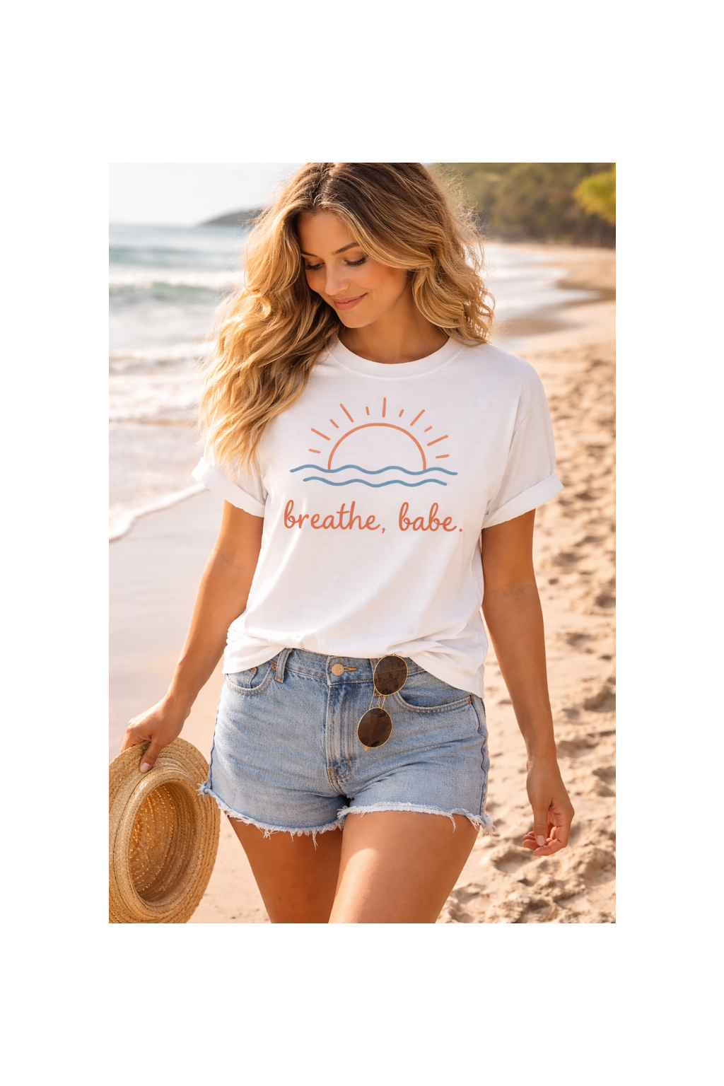 Woman on a beach wearing a white t-shirt with a sun and wave design, and the text 'breathe, babe'.